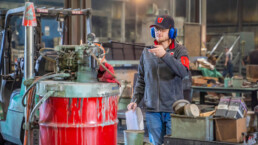 A person wearing university of utah branded clothing and a hat is pointing at a 50 gallon drum inside of what appears to be an industrial space. There is also a fork lift in the midground of the picture.