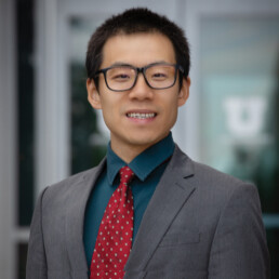 Chauncheng Duan in grey suit and tie wearing glasses and smiling.